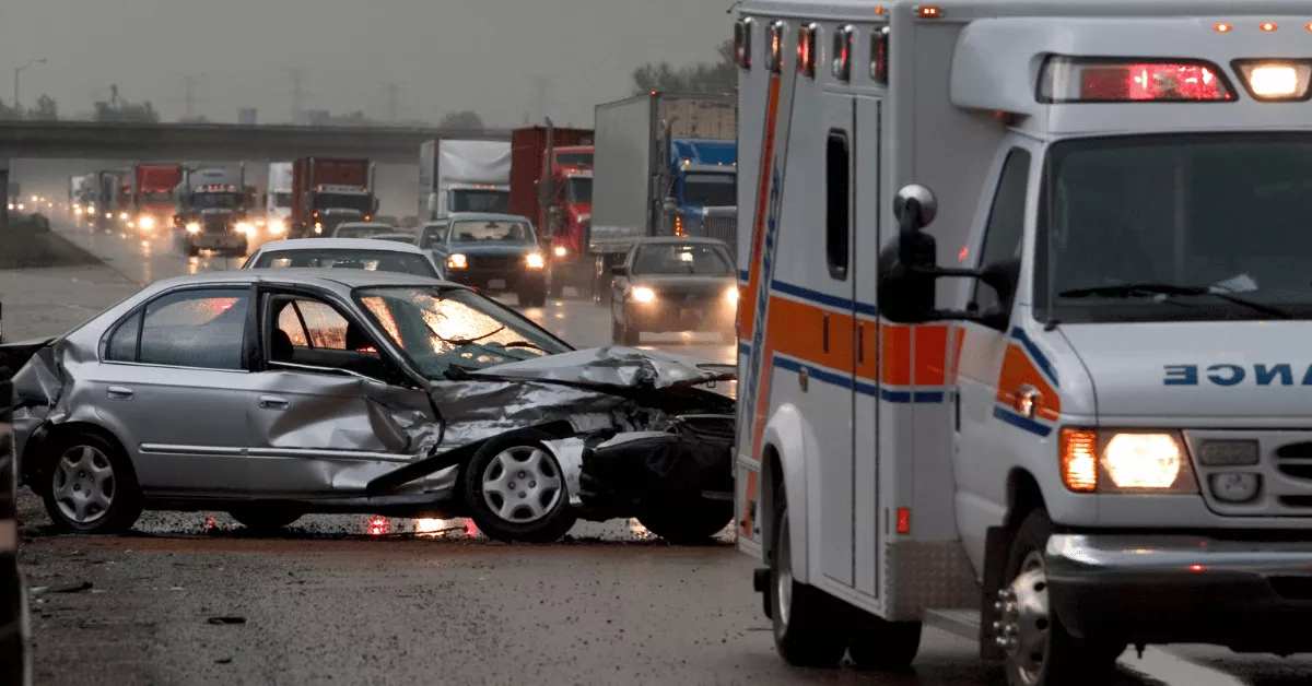 A car accident scene with a mangled silver car, and an emergency ambulance pulled next to the car.